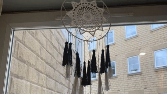A dream catcher hung from a window frame. It is made from string and has a patterned sun design