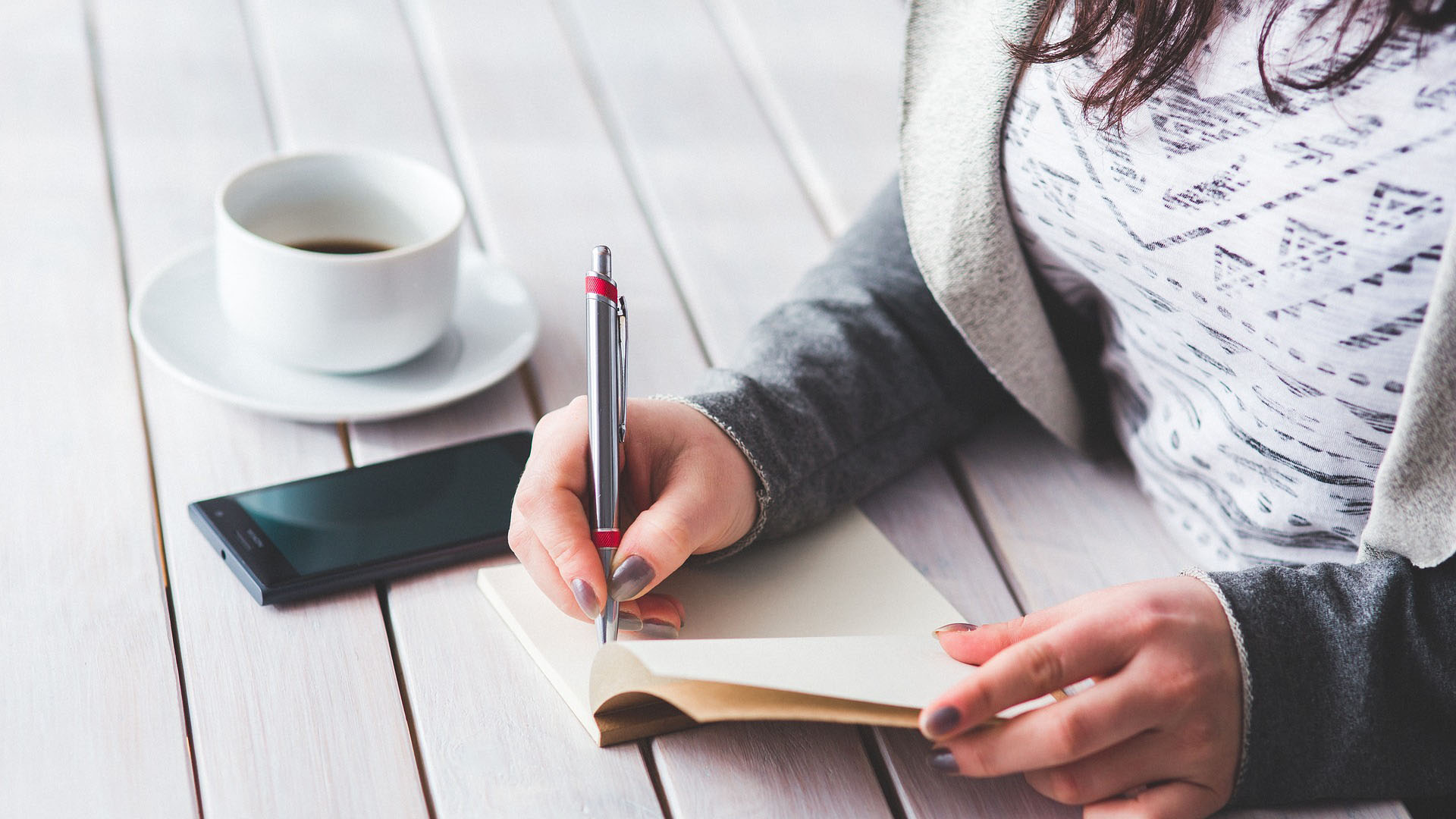 Person writing on a notebook on a desk with a phone and coffee next to them