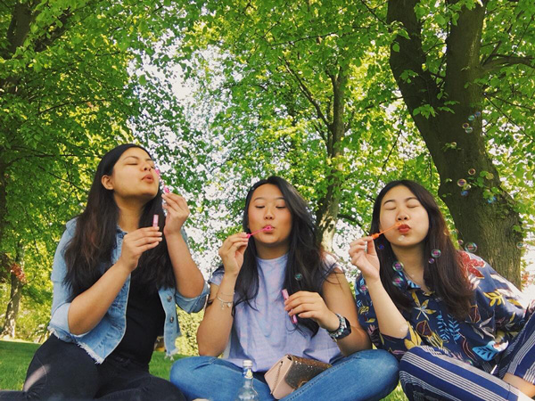 Three Chinese students laying under some trees blowing bubbles