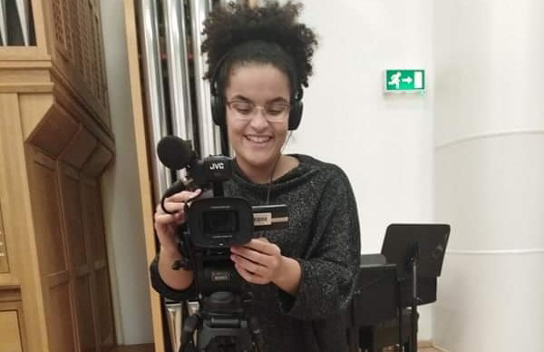 A photo of Kerri operating a camera on a tripod and wearing a pair of headphones.