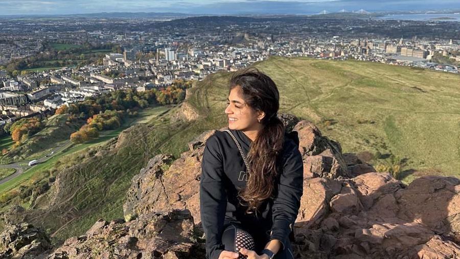 Student Mishti hiking, sat on a stone smiling with the city of Edniburgh in the background