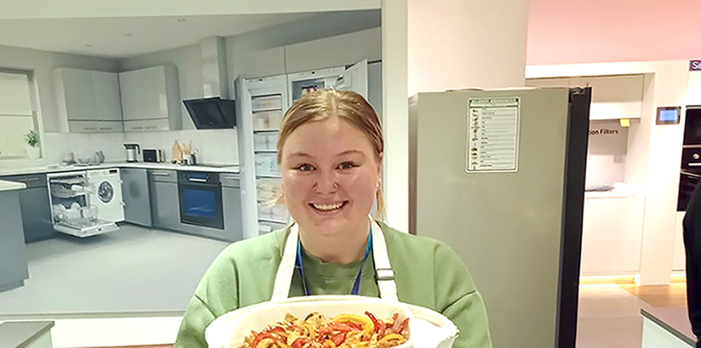 Beth holds up a dish of food in a kitchen while doing some training/team building in a showroom