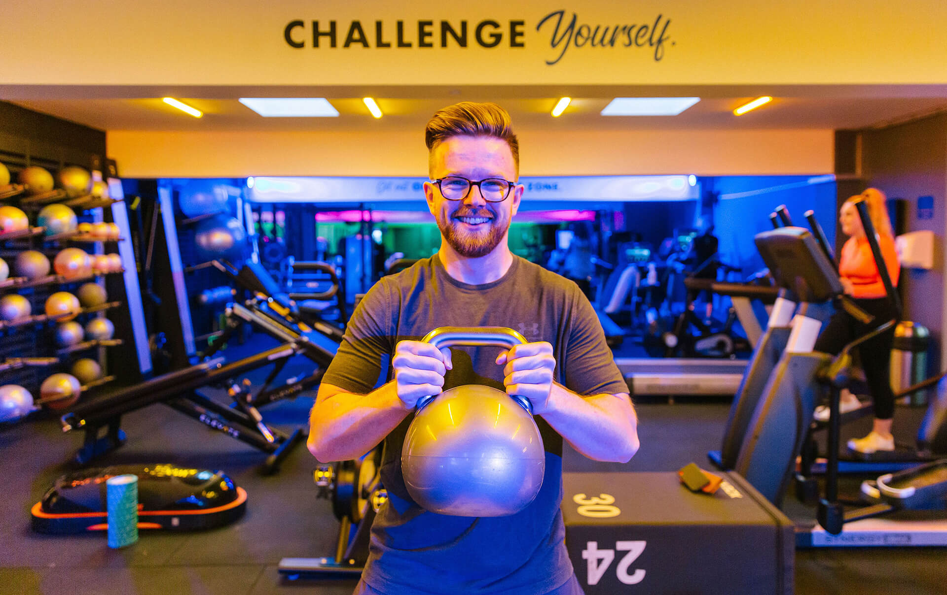 A white male wearing glasses with a hard part fade hair style smiles back at the camera, the background is a well lit gym. Subject is holding a large kettlebell.