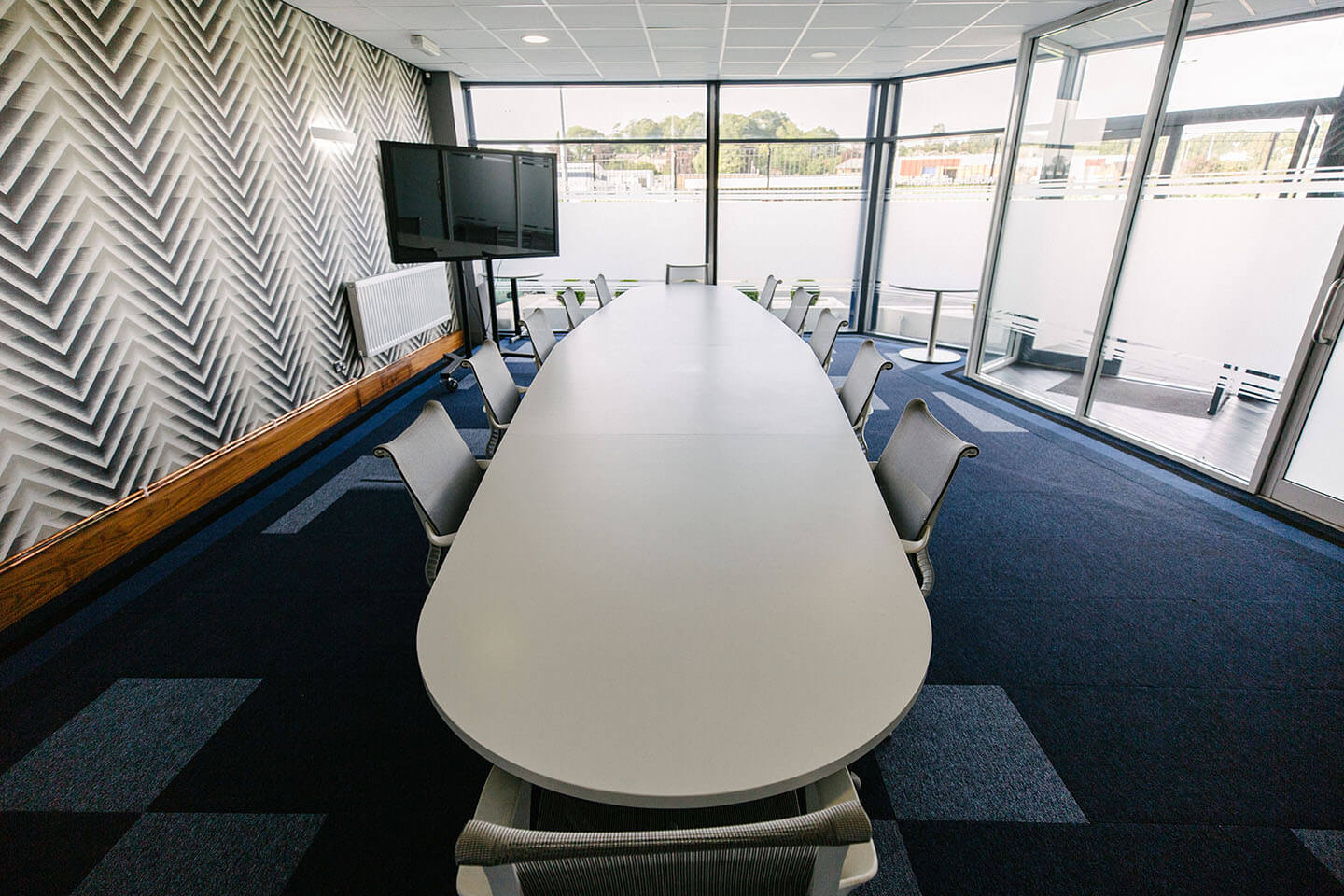 A typical boardroom, a large white table centres the room, blue tiled carpet fills the large room with plenty of natural light.