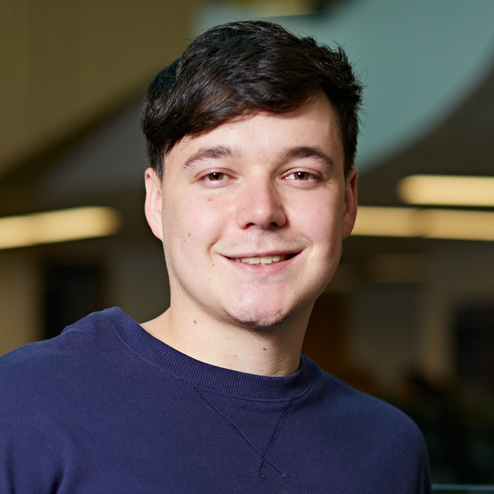 Tom a white male early 20s student smiles back at camera. Tom has hazel brown eyes and dark brown hair and wears a dark blue crewneck jumper