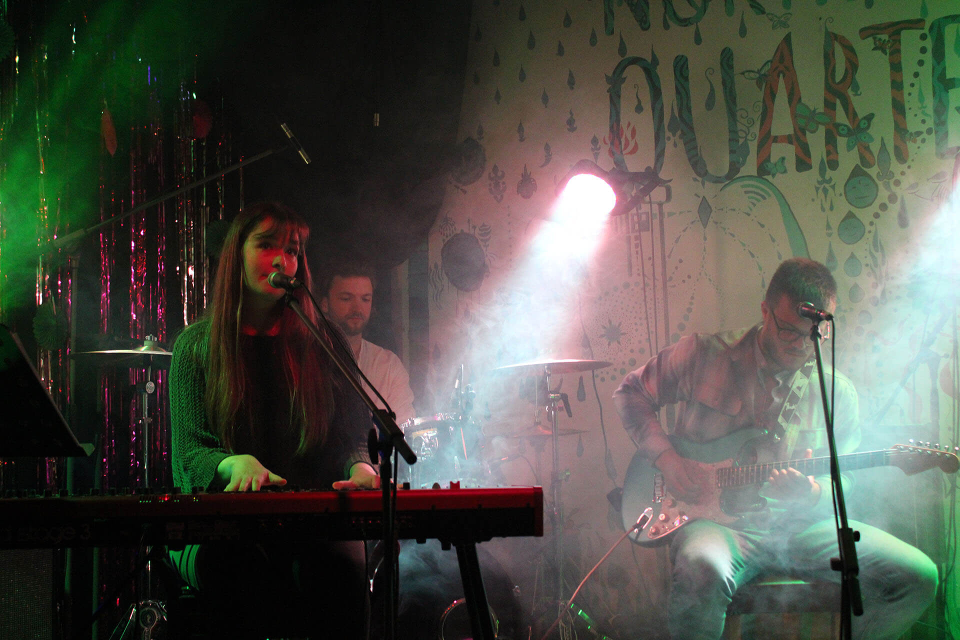 A woman plays a keyboard on stage, next to a man playing a guitar, while a man plays drums in the background. Beside them wording on the wall says Northern Quarter