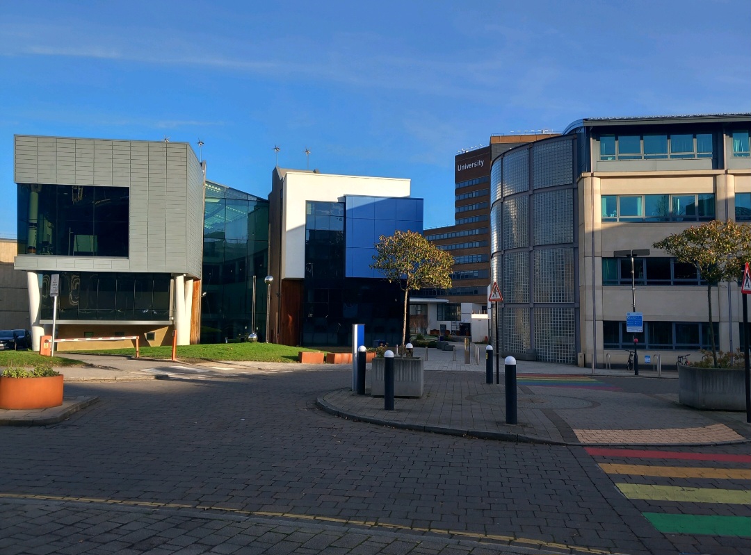 University of Huddersfield campus entrance with multiple contemporary buildings, a circular paved area with benches, and a small tree in a planter. The architecture features glass and metal facades under a clear blue sky.