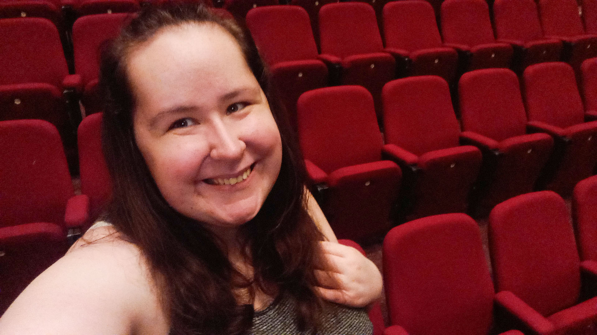 Smiling person taking a selfie in a theatre with multiple rows of empty red seats in the background.