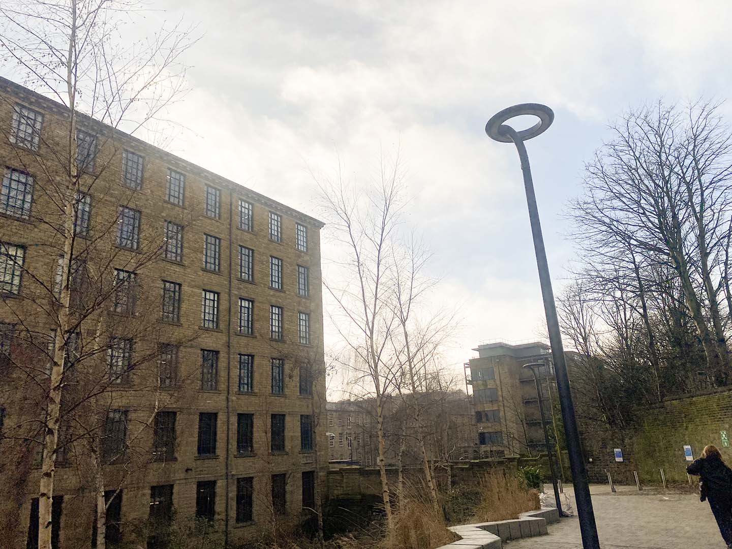 View of Huddersfield's Sparck Jones, a historical mill building, next to leafless trees and a modern street lamp, under a cloudy sky.
