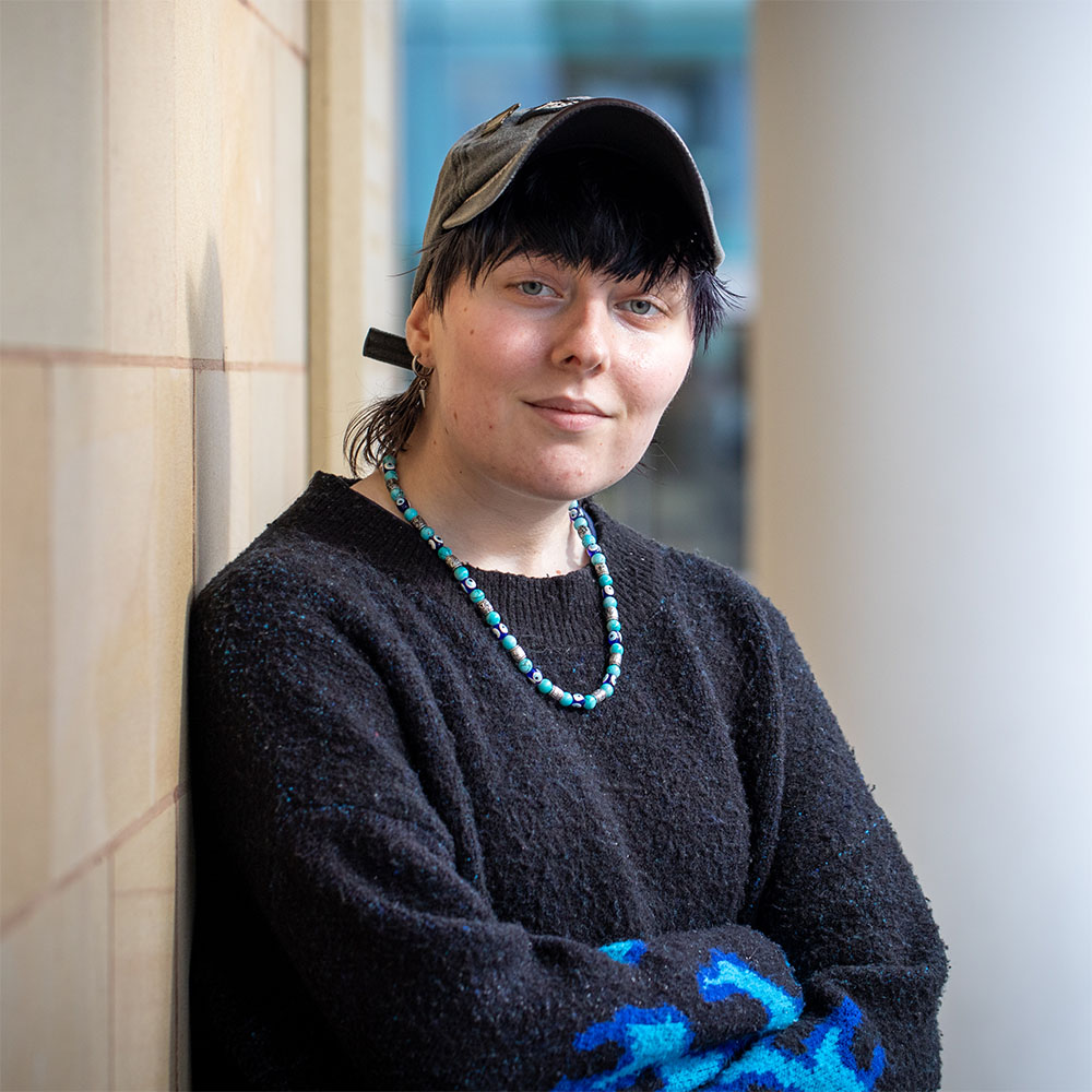 A person wearing a black sweater and a baseball cap smiles at the camera, with a tan brick wall in the background. The person is adorned with a blue and green beaded necklace.