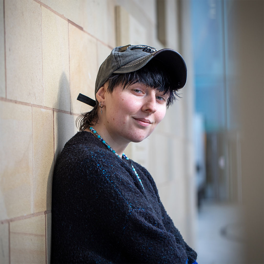 Person wearing a baseball cap and a black sweater, smiling at the camera in front of a beige wall.