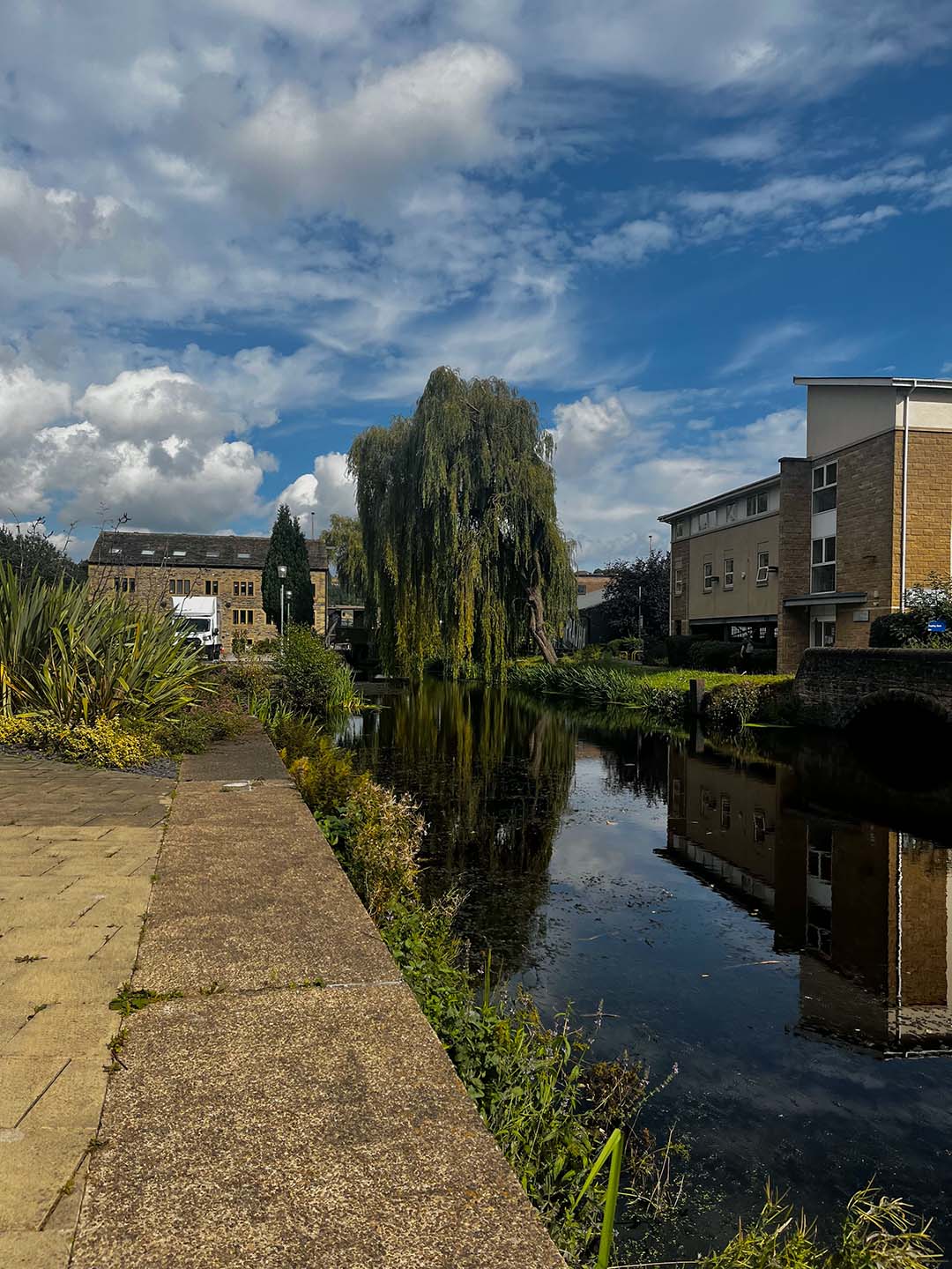 A scenic view along a the Huddersfield narrow canal at the University of Huddersfield with a weeping willow on the opposite bank. Old and modern buildings flank the riverbanks under a partly cloudy sky.