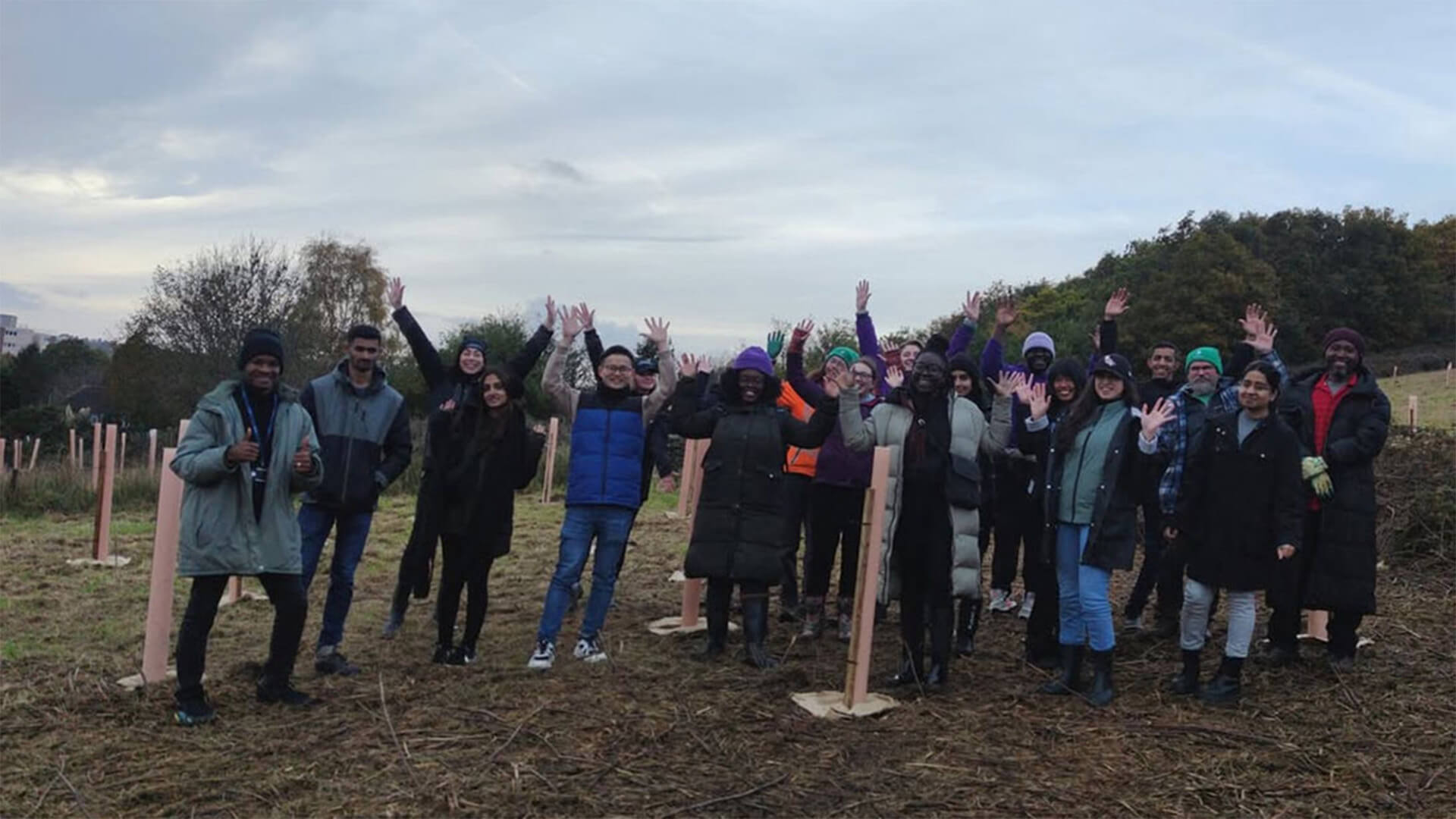 A group of happy individuals celebrating, raising their hands in the air, while standing in a field with wooden stakes placed in the ground. They are dressed warmly and the background features overcast skies and distant hills.