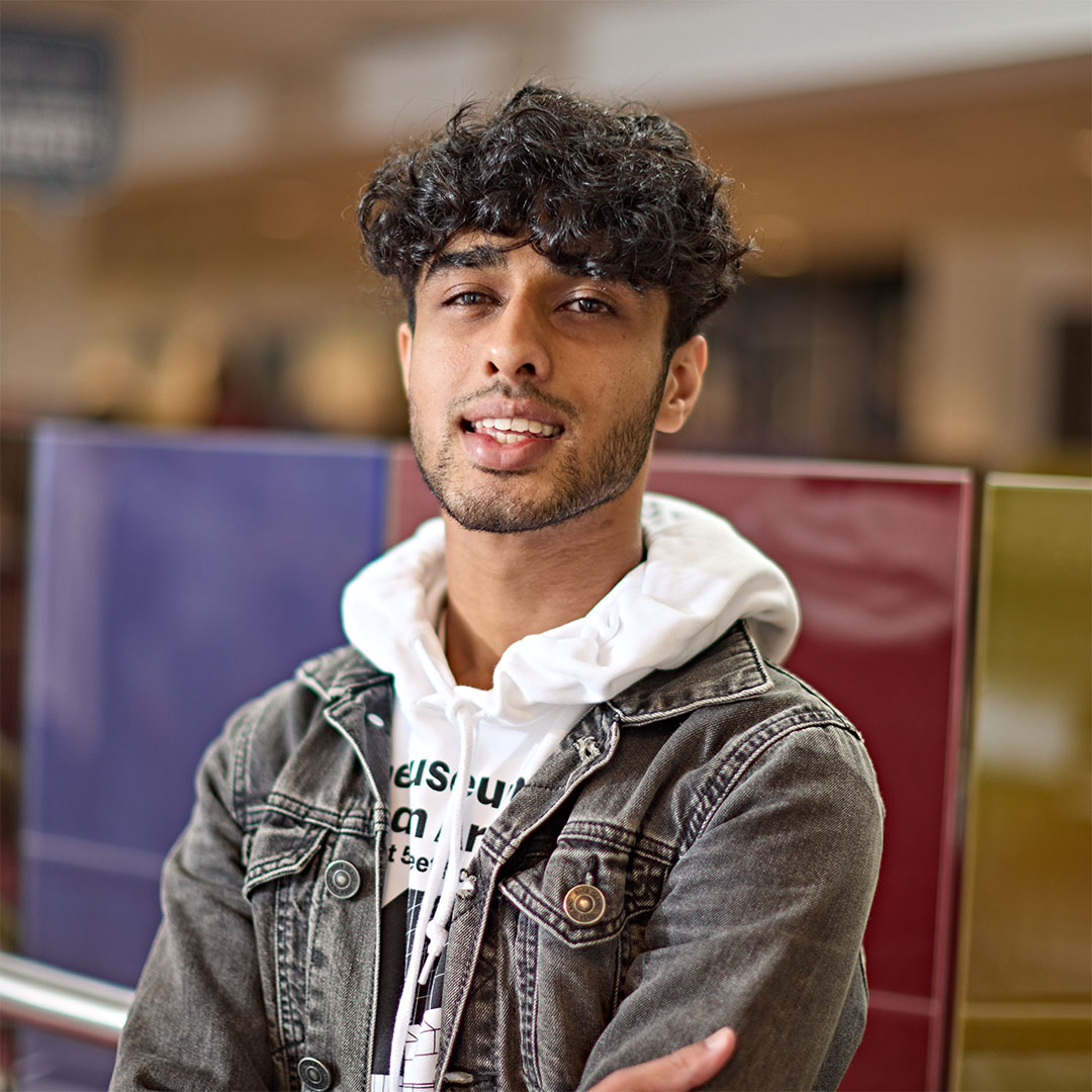 Portrait of a smiling Ismaeel, with curly hair, wearing a denim jacket over a hoodie, in the Students' Union at the University of Huddersfield.
