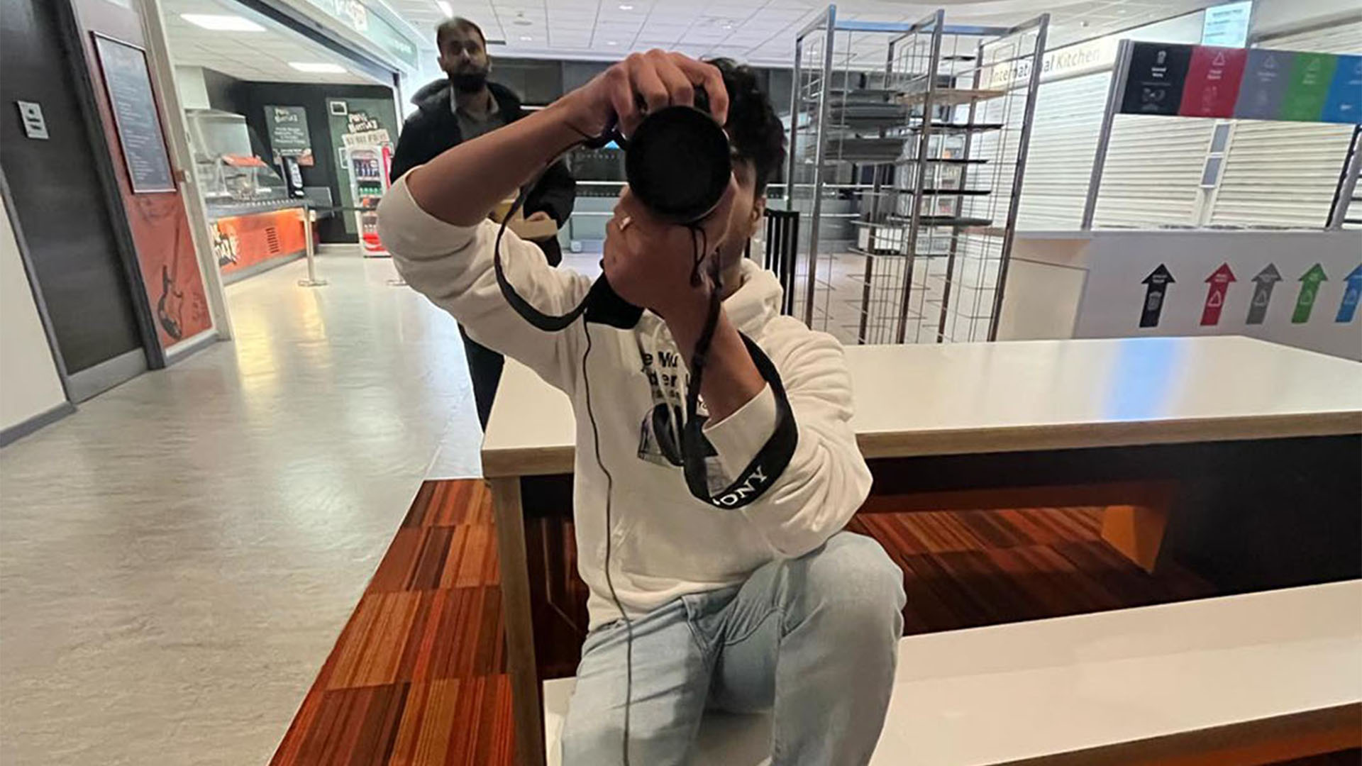 Person sitting on a bench using a camera and lens in a public indoor space.
