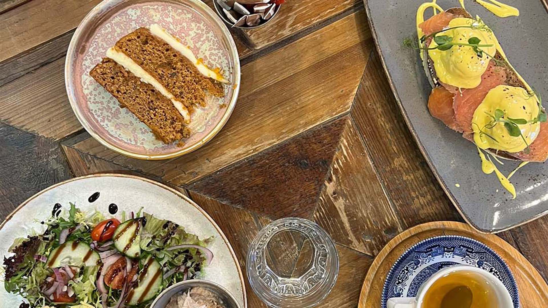 Overhead view of a breakfast spread on a wooden table, featuring dishes including eggs benedict, toast with spread, a salad with mixed greens, and a bowl of rice with sides. Accompanied by two cups of coffee and a drink in a clear glass. Various condiments and utensils are also on the table.