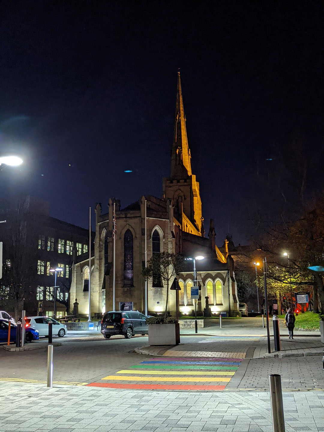Night view of St. Paul's Church with illuminated windows and a tall spire, located beside a street with a rainbow crosswalk.