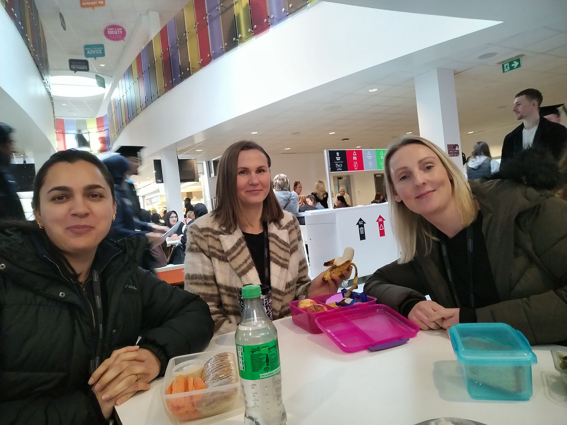 Three individuals sitting at a table in a busy cafeteria, sharing a meal, with various drinks and tupperware on the table.