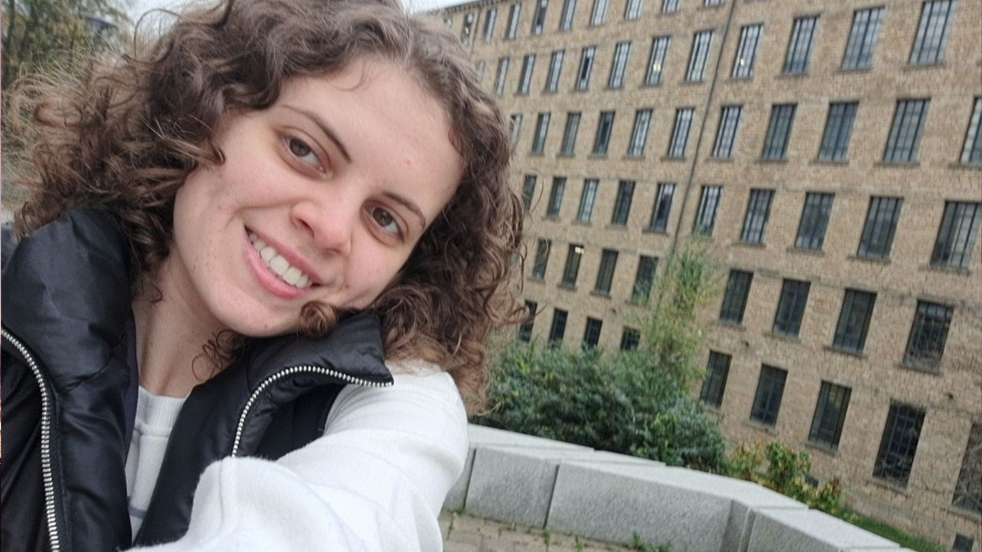 A smiling student taking a selfie in front of a mill building