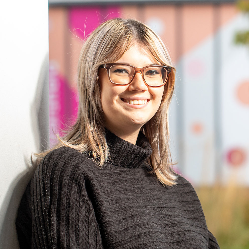 Person smiling and wearing glasses, leaning against a wall outdoors. The individual has shoulder-length hair and is dressed in a black turtleneck sweater. The background includes soft pink tones and blurred circular designs.