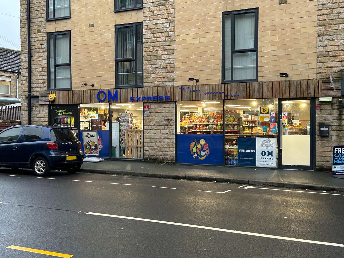 Exterior view of a DM Express convenience store with visible logo and colorful product displays inside, located in a stone building on a street. A parked car is visible in front.