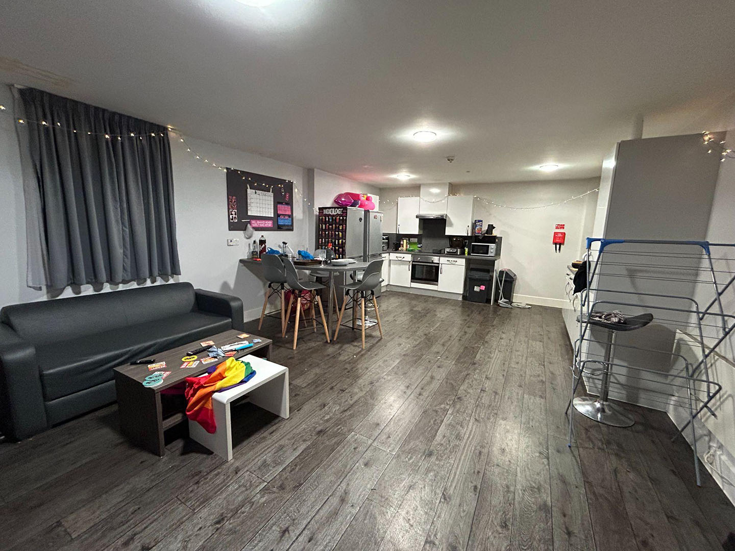 Interior view of a modern living space with a kitchen area, dining table, sofa, and coffee table. Decorations include fairy lights and a wall-mounted neon sign.