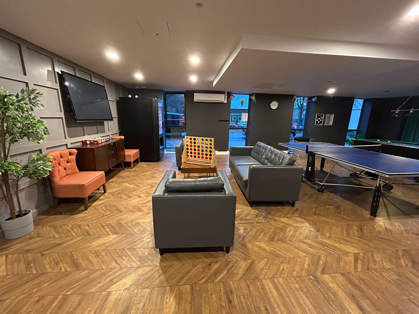 A spacious recreation room featuring a ping pong table, a foosball table, a large TV, and a seating area with a sofa and armchairs. The room has wooden flooring and is well-lit.