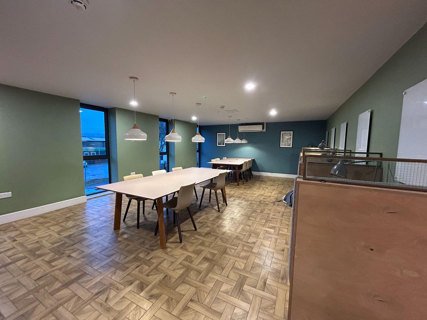 Castings study area. Interior of a modern room with green walls featuring a wooden floor, multiple tables, and pendant lights, providing a view of an outdoor body of water through large windows.