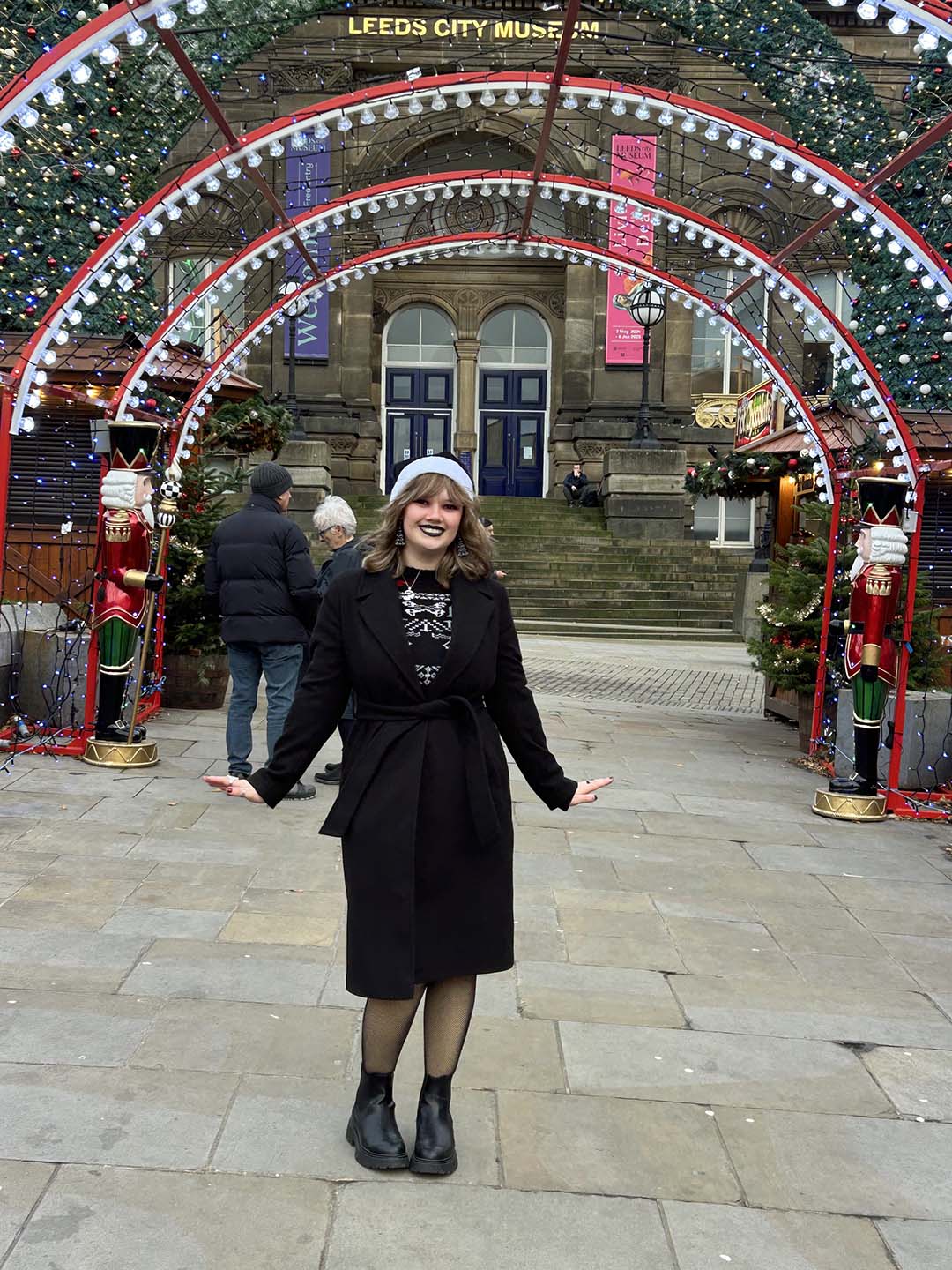 Maisy standing joyfully at Leeds Christmas Market in front of Leeds City Museum, which is decorated with festive lights and Christmas decorations.