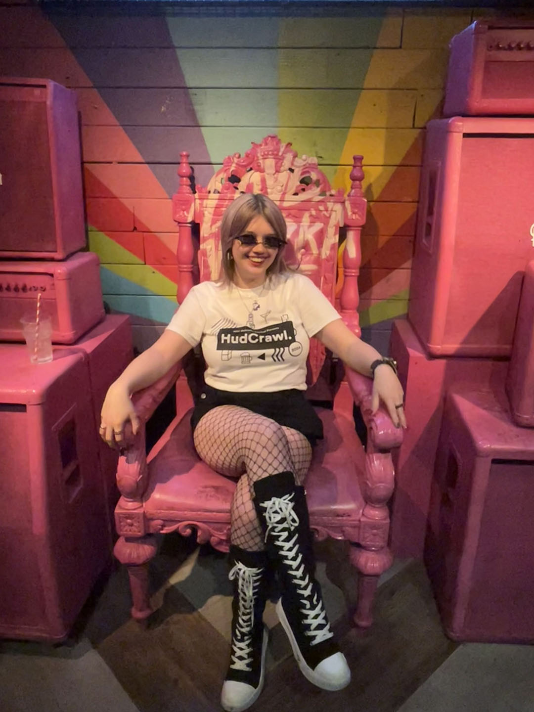Maisy wearing a 'HudCrawl' t-shirt, sunglasses, and fishnet stockings, sitting on a large pink throne surrounded by pink washing machines in a playful setting.