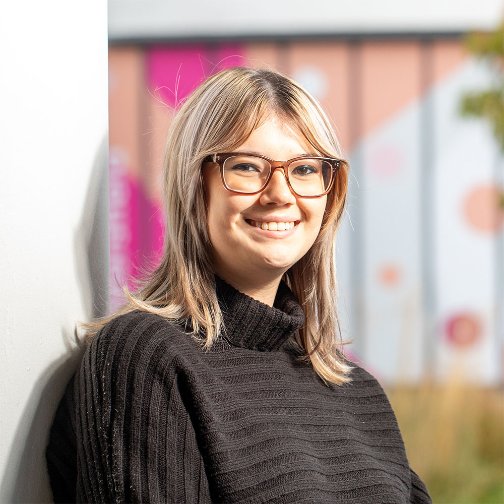 Portrait of a smiling person with glasses, wearing a black sweater, standing against a blurred urban background.