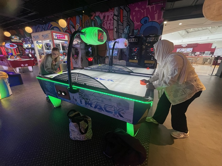 Two people play air hockey in an arcade with a graffiti background and colourful lighting