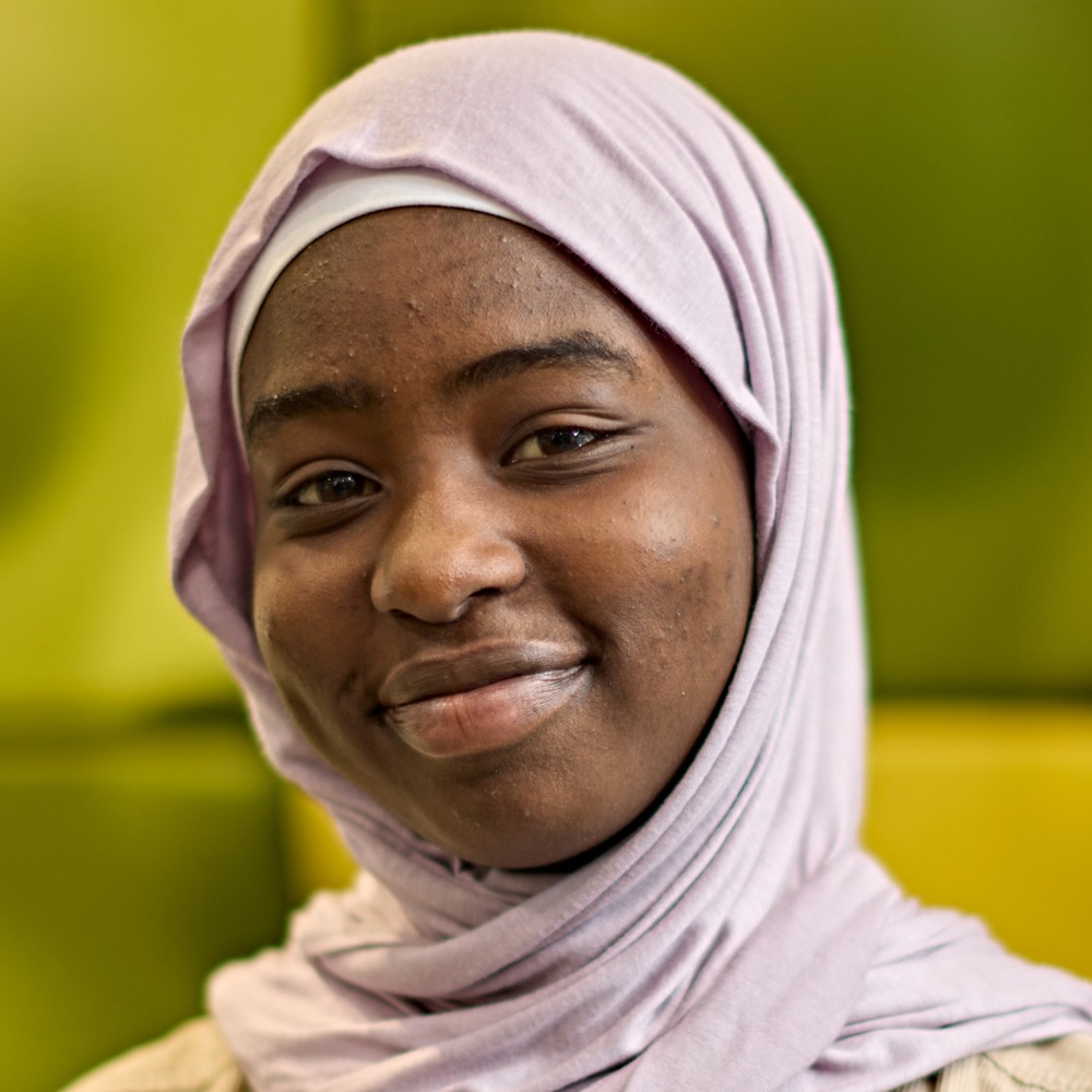 A student wearing a pink hijab smiles towards the camera, against a bright green background