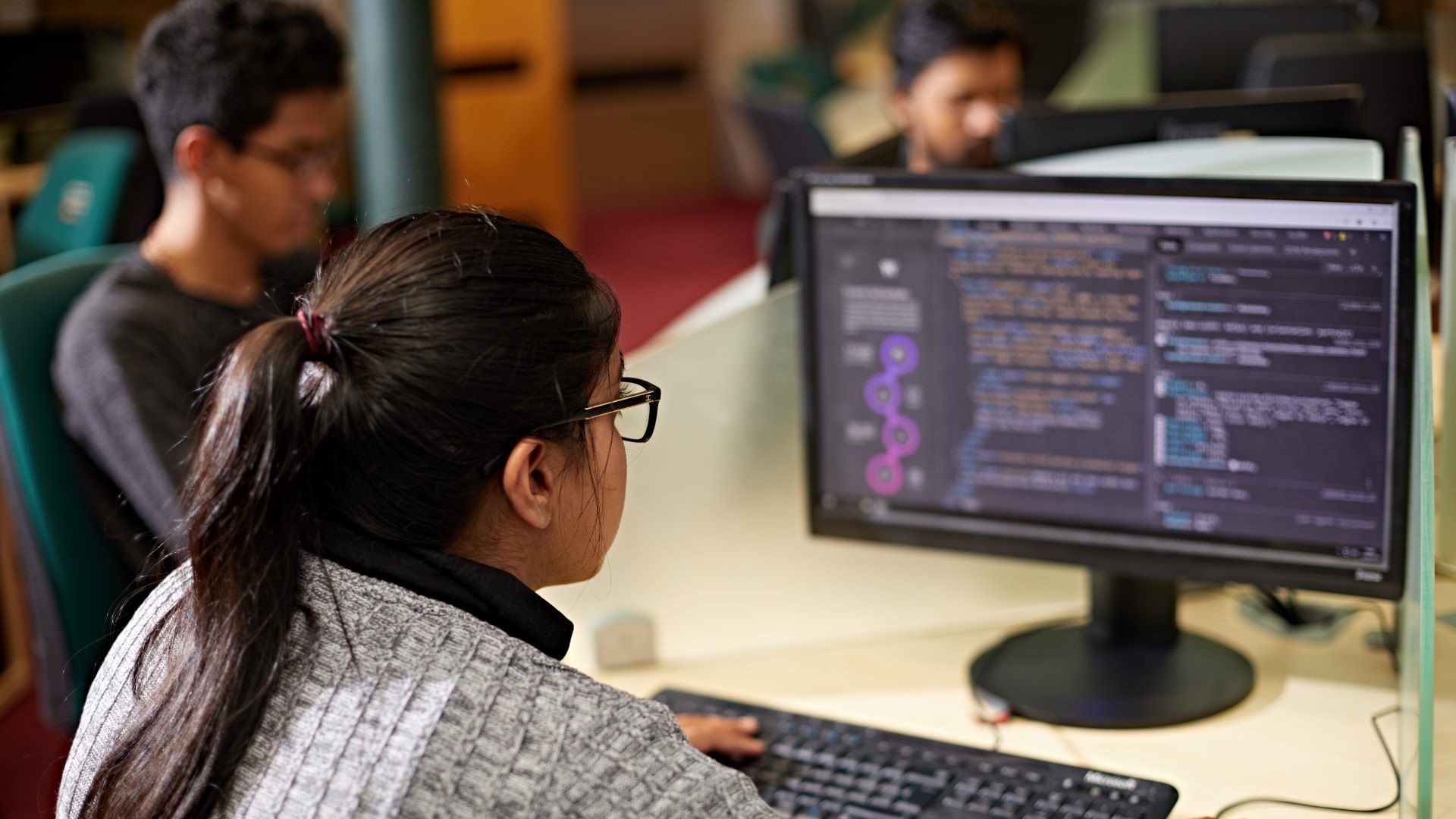 Person using a computer with code on the screen in an office setting, with another individual visible in the background.