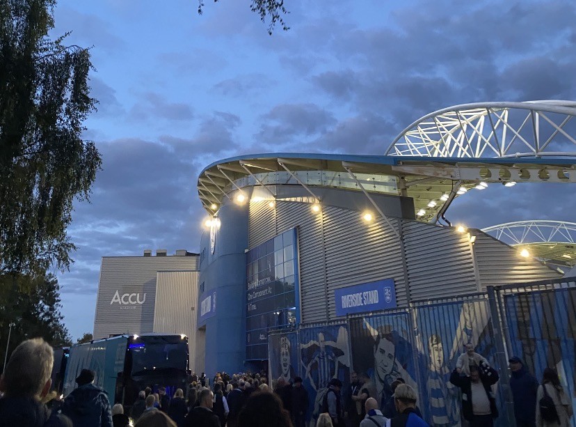 A football stadium at dusk with a crowd of fans walking towards the stadium