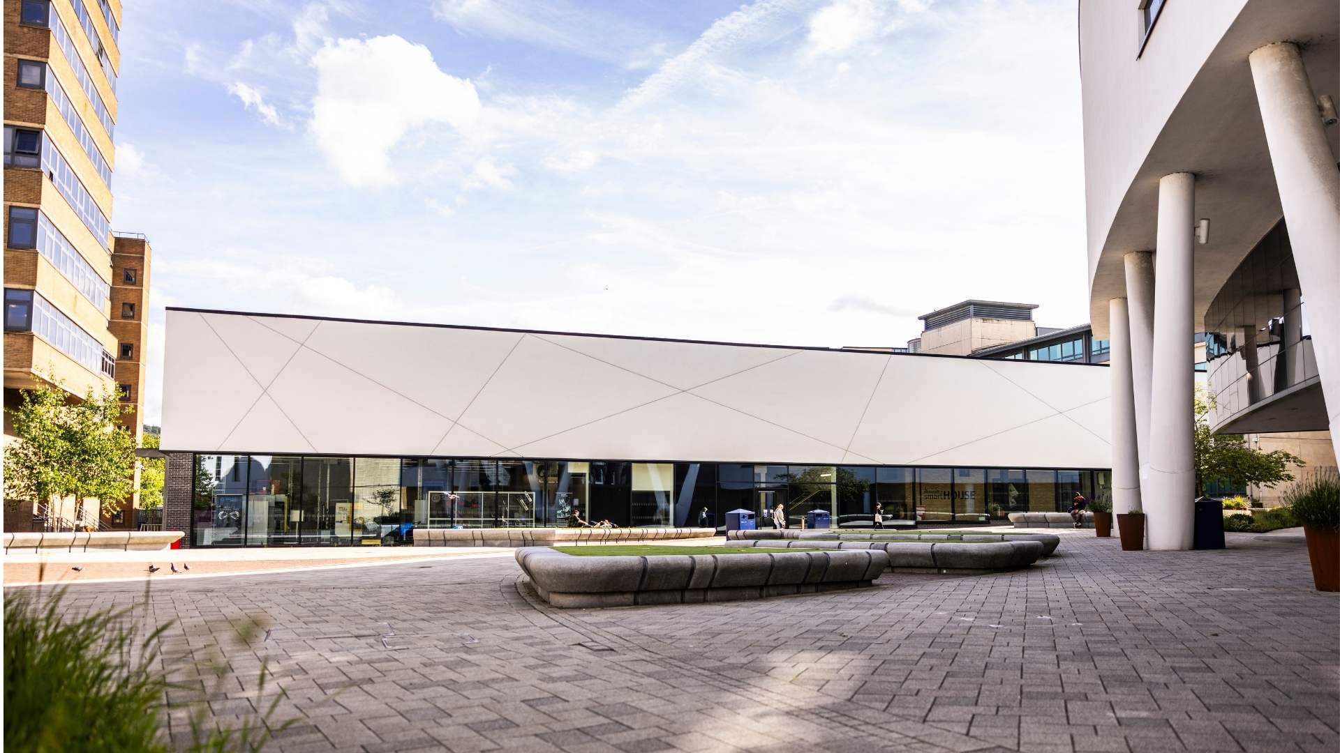 Modern building with geometric facade and glass windows, surrounded by a paved plaza. Urban area with trees, benches, and additional structures nearby under a blue sky.