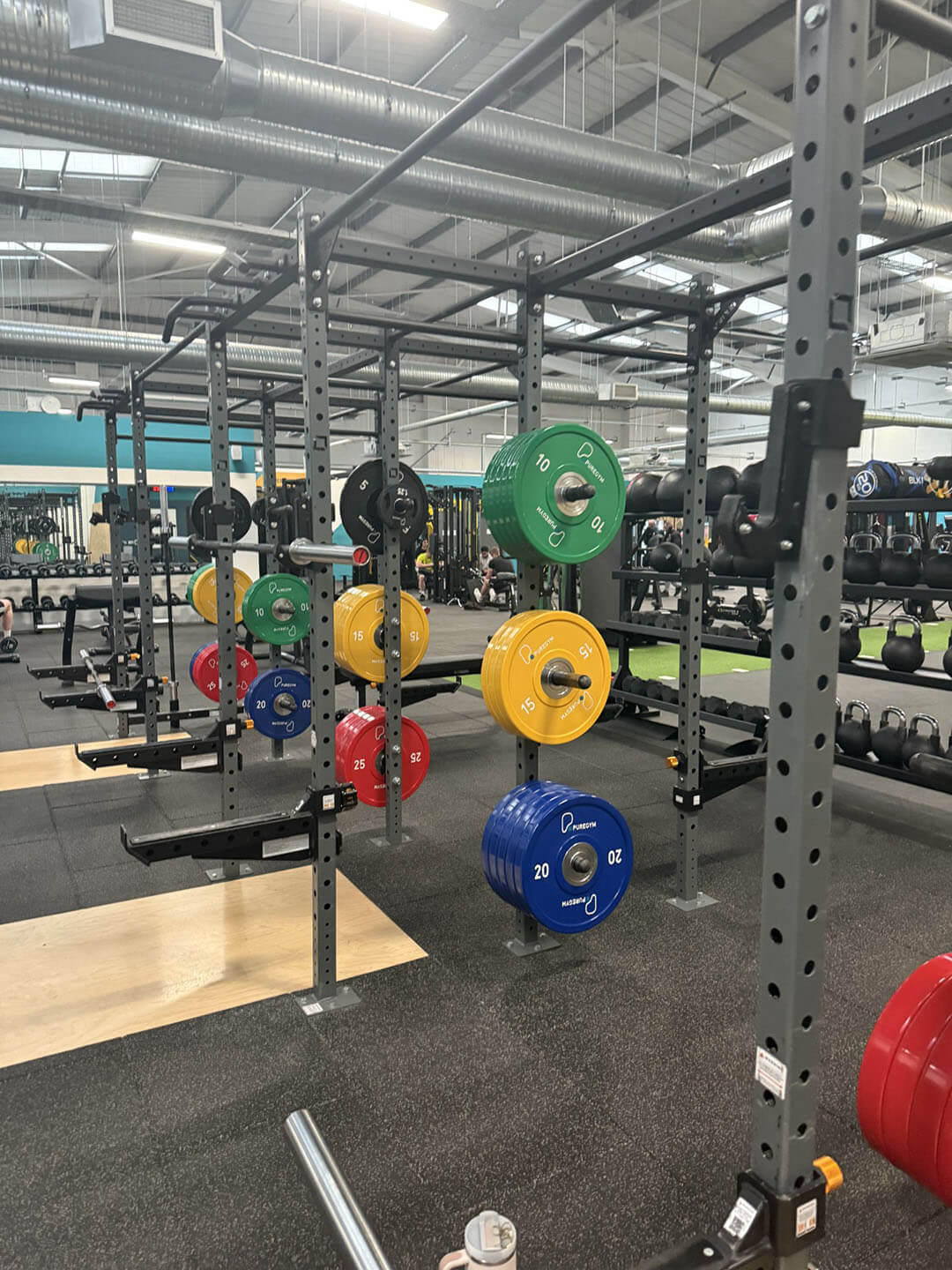 A gym with several squat racks and weightlifting platforms. Brightly colored weight plates are organized on racks, including blue, red, yellow, and green plates. The background shows other gym equipment like kettlebells. The setting is spacious and well-lit with industrial-style ceilings.