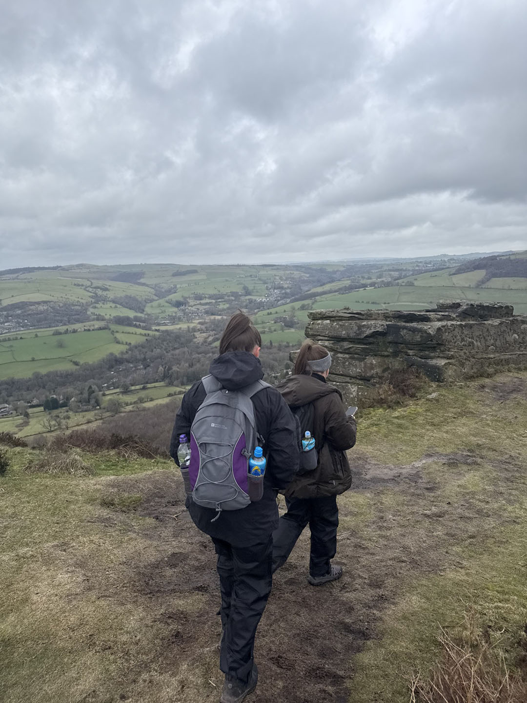 Two people walking along a path with backpacks containing water bottles. They are overlooking a scenic, hilly landscape with fields and distant valleys under a cloudy sky.
