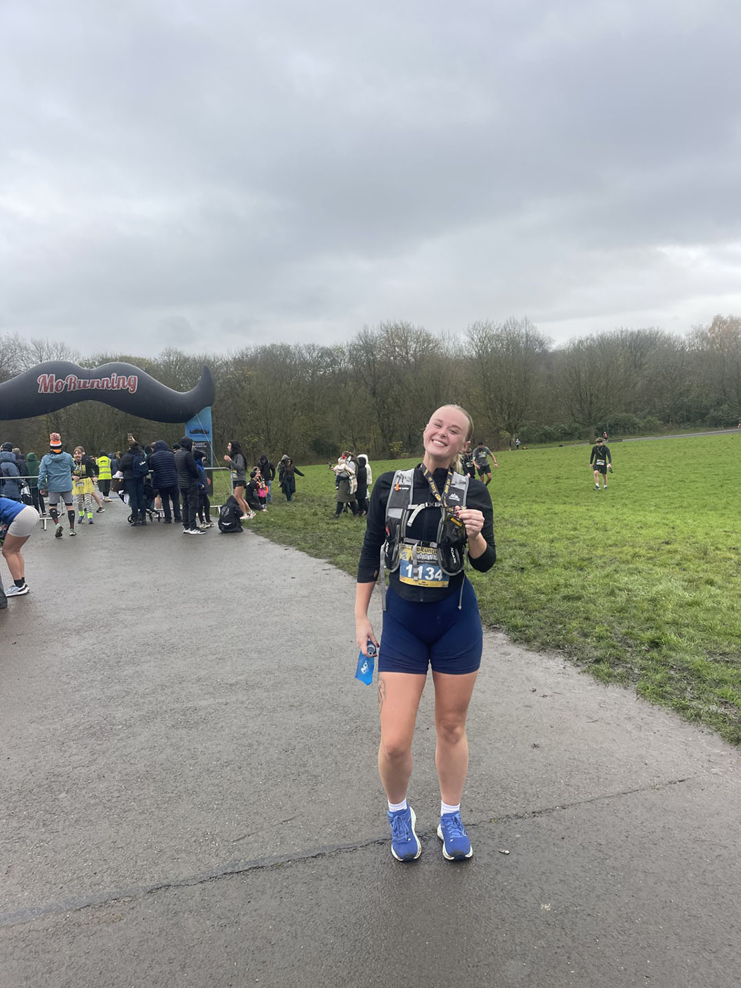 A person stands on a paved path holding a medal, wearing running gear. In the background, there are people gathered near an inflatable archway labeled