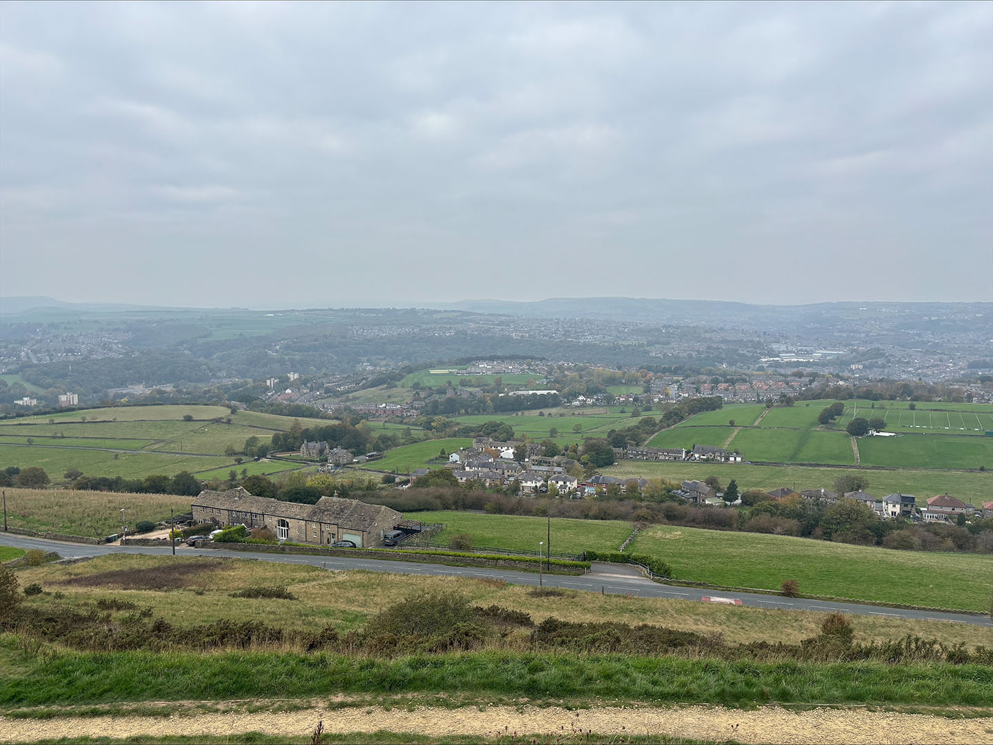 Panoramic view of a landscape featuring a sprawling green valley with scattered buildings and a road, under a cloudy sky.