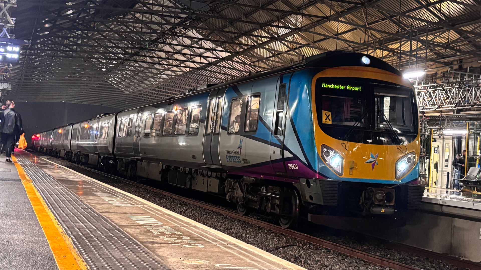 A Northern Rail train at a station, labeled for Manchester Airport, parked under an expansive metal roof structure at night.