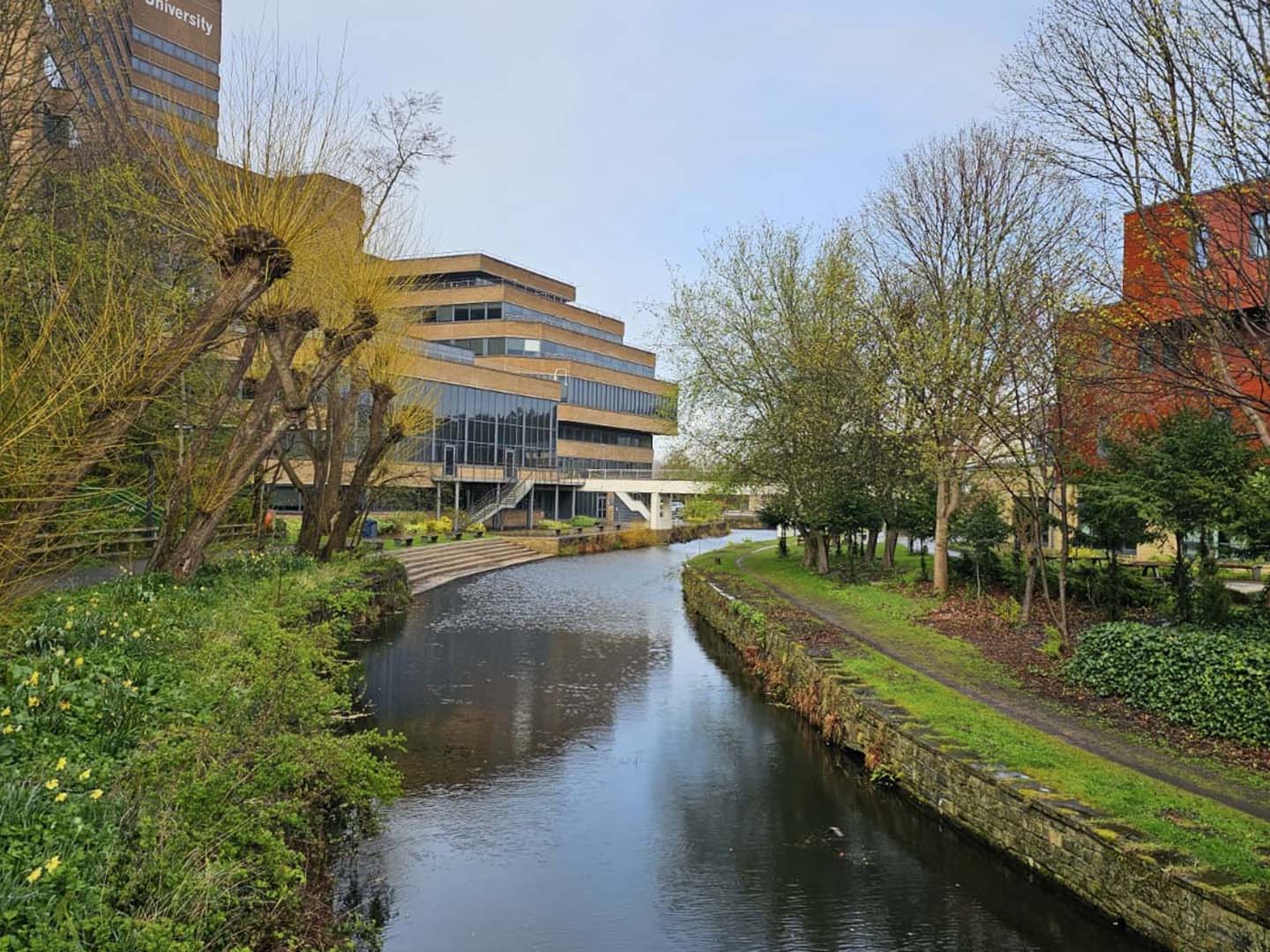 A serene canal surrounded by green trees and bushes runs through an urban area with modern buildings. A tall building with the word