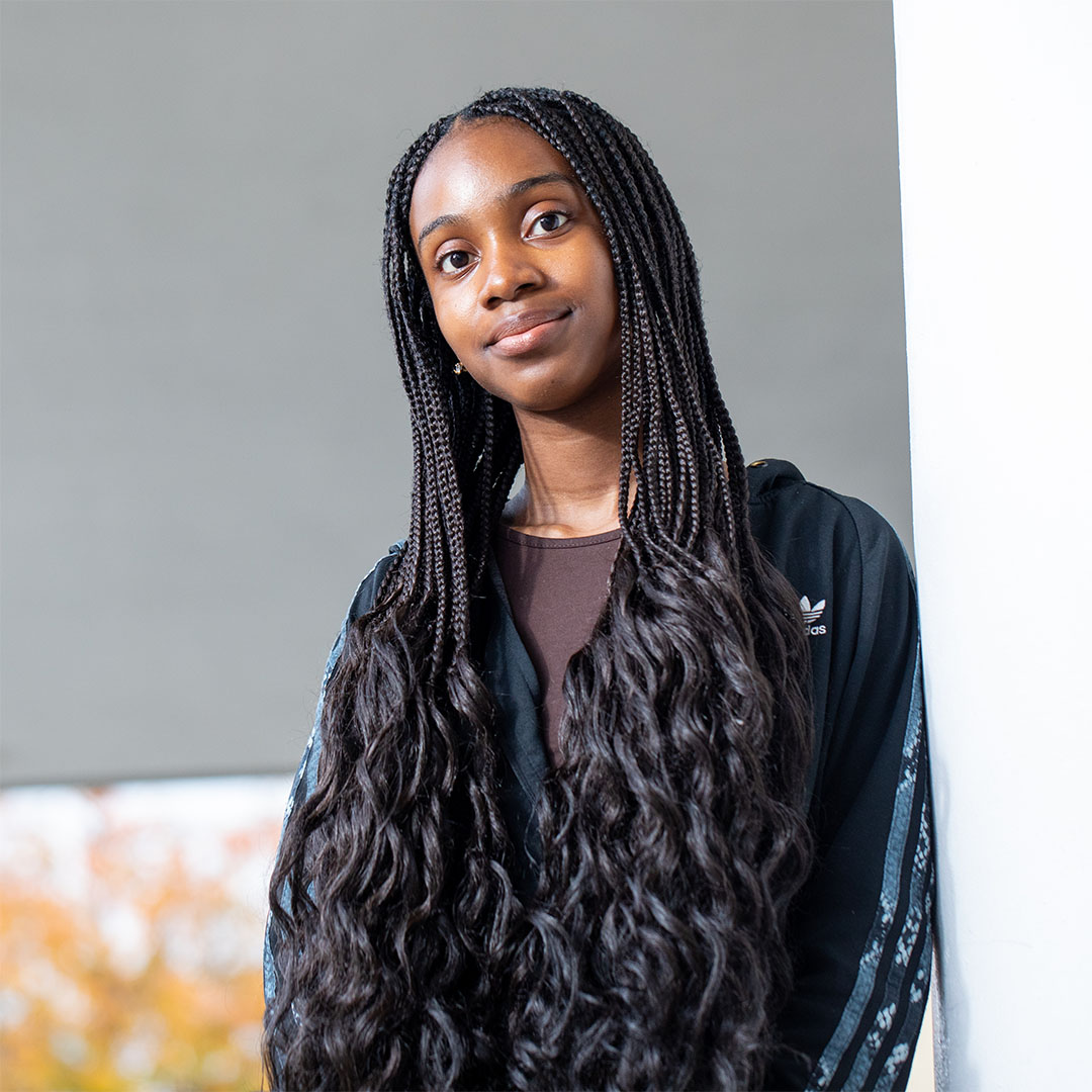 Tofunmi smiling back at the camera. She wears a dark brown tee, a black adidas hoodie and has her hair braided with the ends curled / french curl braids.
