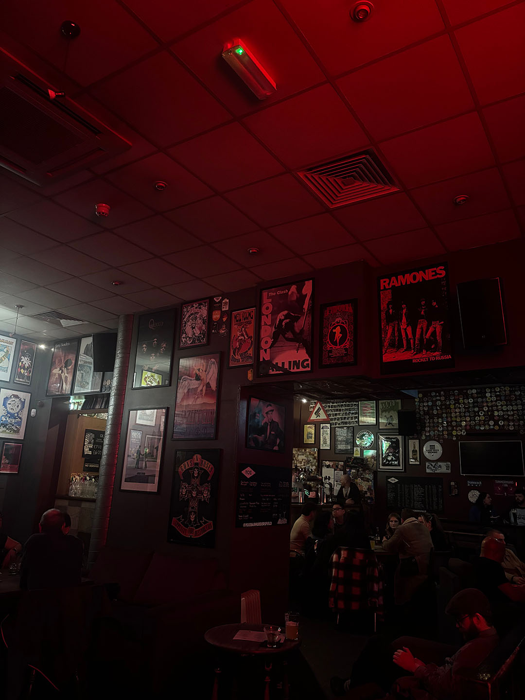 Interior of a Vox bar in Huddersfield, a vibrant bar with red lighting, showcasing walls adorned with framed rock band posters, including the Ramones. People are seated, enjoying the ambiance.