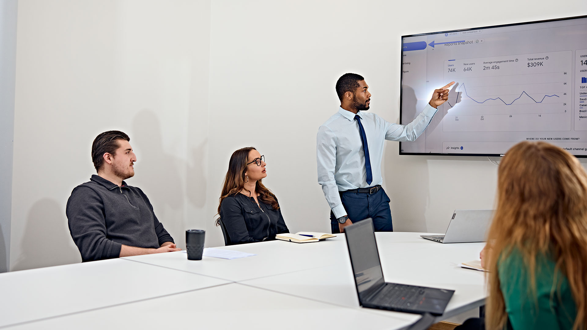 A person in a shirt and tie, leading a business meeting with three other people.