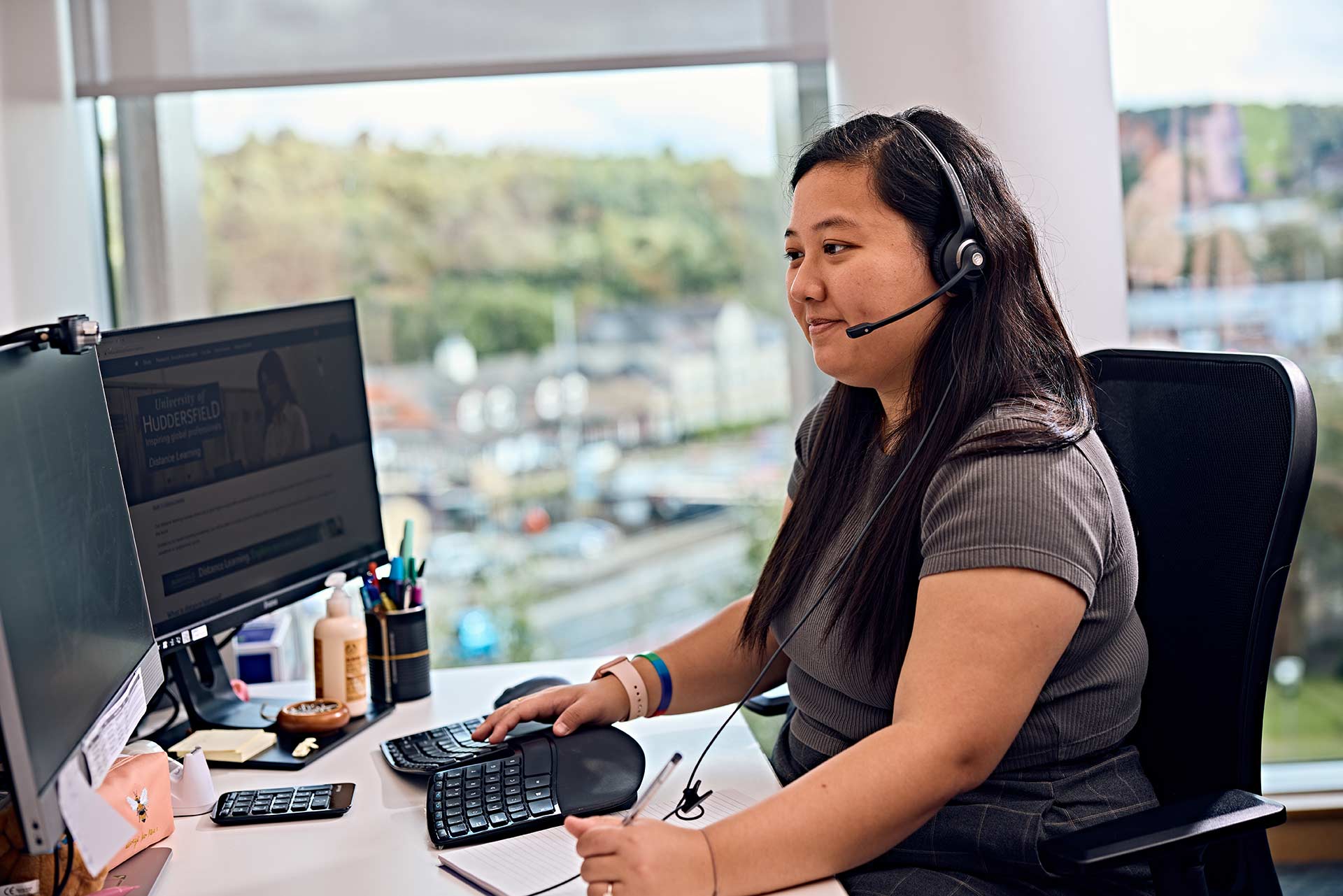 A young adult wearing a headset, working in an office on a computer.