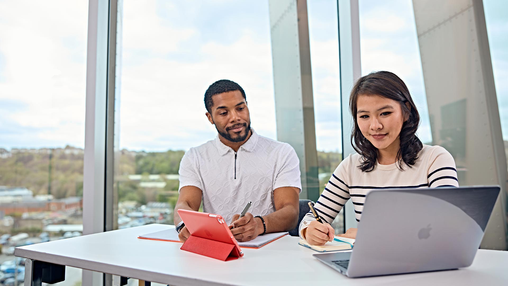 Two people sat at a desk working together, one has a laptop, one has a tablet device.
