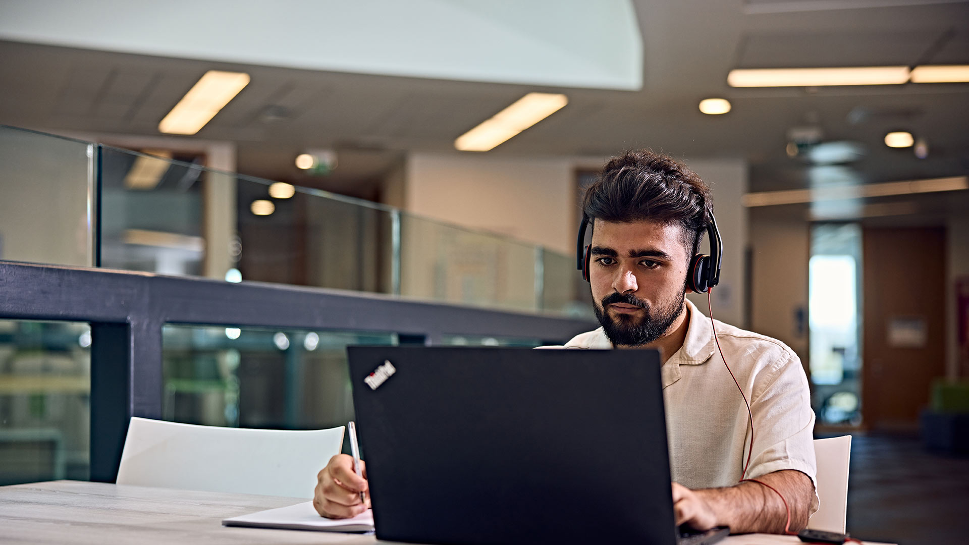 A student with a headset on, they are writing notes whilst looking at their laptop.
