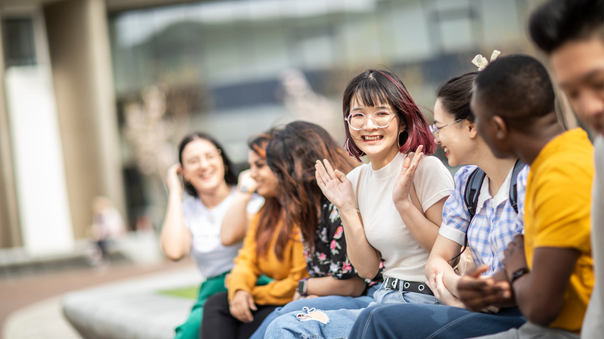 A group of postgraduate international students talking and laughing on a bench outside main campus buildings